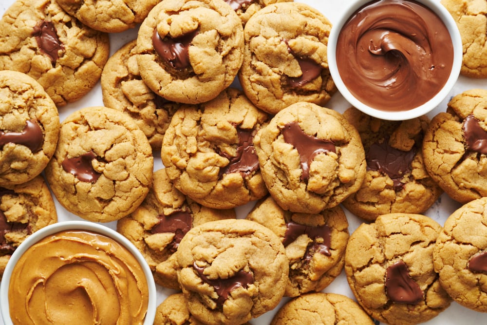 Een volledige batch pindakaas-Nutella koekjes, vers gebakken, met schaaltjes romige pindakaas en Nutella in de buurt voor extra verwennerij.